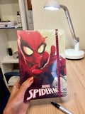 Marvel Spider-Man notebook held by a hand on a desk with a lamp and pen in the background