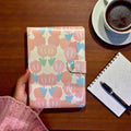 Floral patterned notebook held by a hand on a wooden table with a cup of coffee and pen.