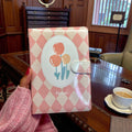 Pink checkered notebook with floral design held by a hand in a room with wooden furniture.